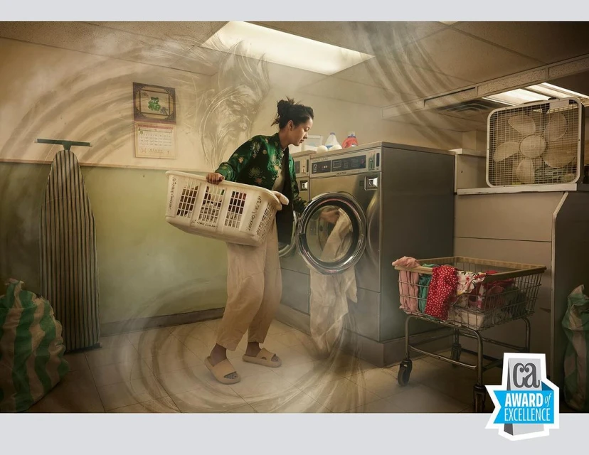 Woman loads laundry into a vintage washing machine in a public laundromat with laundry carts, an ironing board, and a box fan visible.