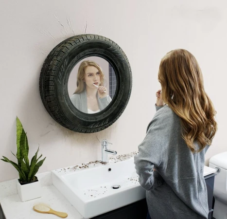 Woman in grey sweater brushes teeth in bathroom; mirror framed with black tire hangs on wall above sink with cluttered countertop.