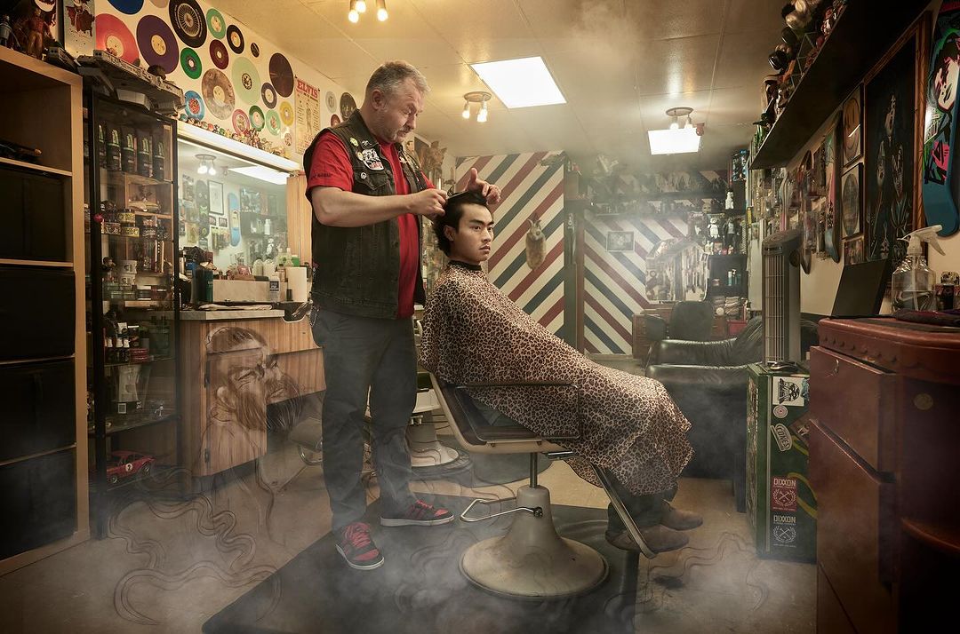Male barber cuts customer's hair in a vintage-style barber shop with record-covered walls and traditional barbering equipment under bright lights.