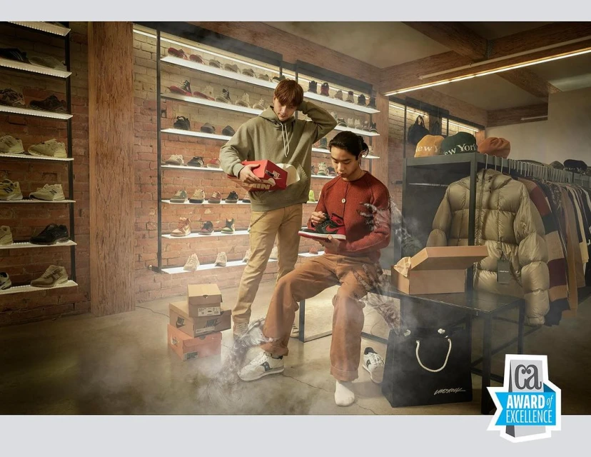 Two stylish men examine sneakers in a well-lit boutique, surrounded by shelves of shoes, clothing racks, and a smokey haze on the floor.