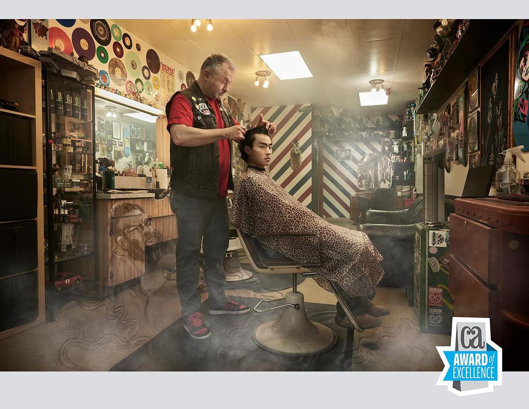A barber cuts a young man's hair in a vintage-style barbershop. The shop features records, vintage decor, and atmospheric lighting.