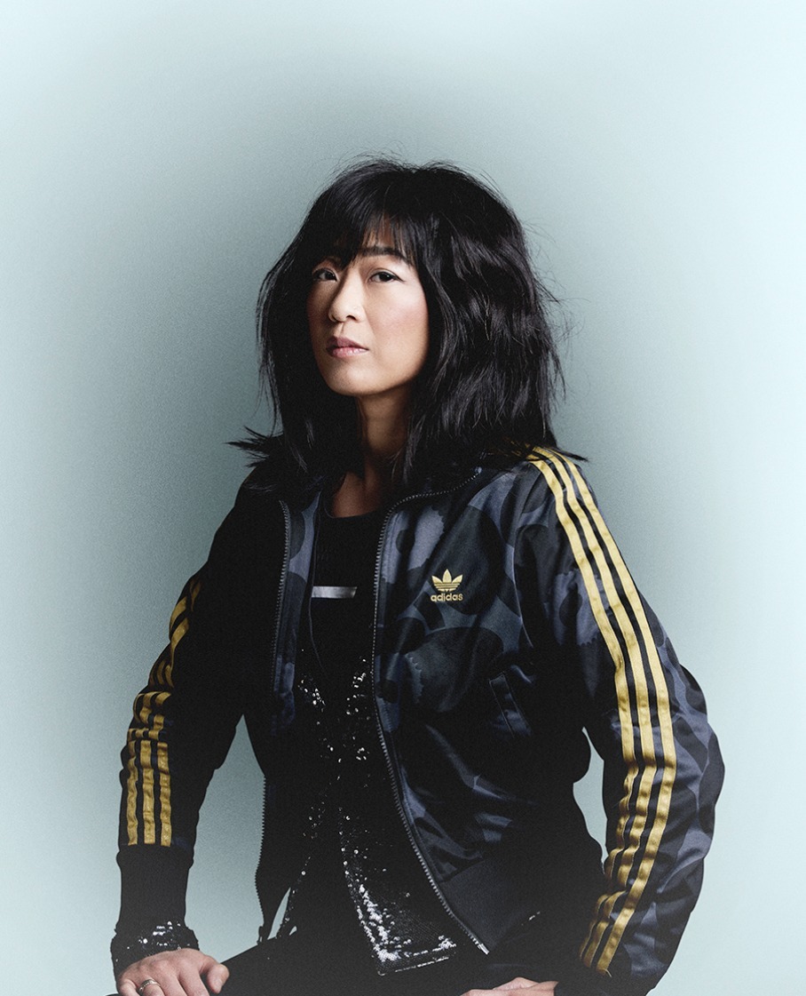 Portrait of an adult woman with dark hair wearing a black jacket with gold stripes, posing against a light blue background.