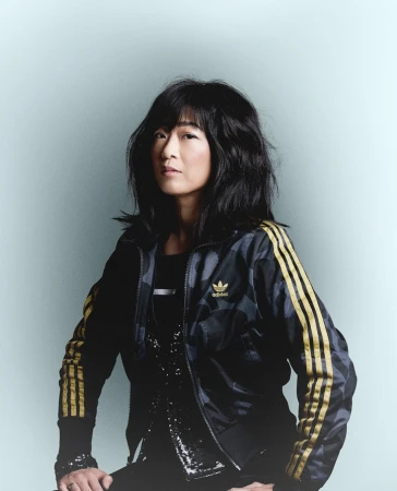 Portrait of a woman with dark hair wearing a patterned track jacket with gold stripes against a pale blue backdrop.