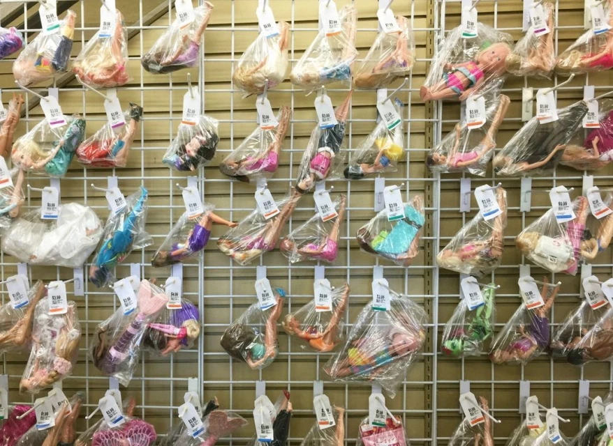 Display of dolls packaged in clear plastic bags and hanging on a metal grid, with price tags, in an indoor retail environment.