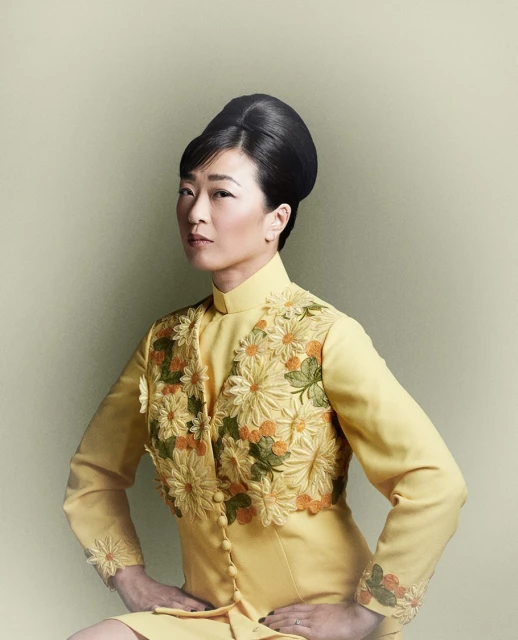 Elegant Asian woman in yellow floral dress poses with hands on hips in a studio portrait. Her dark hair is styled in a beehive updo.