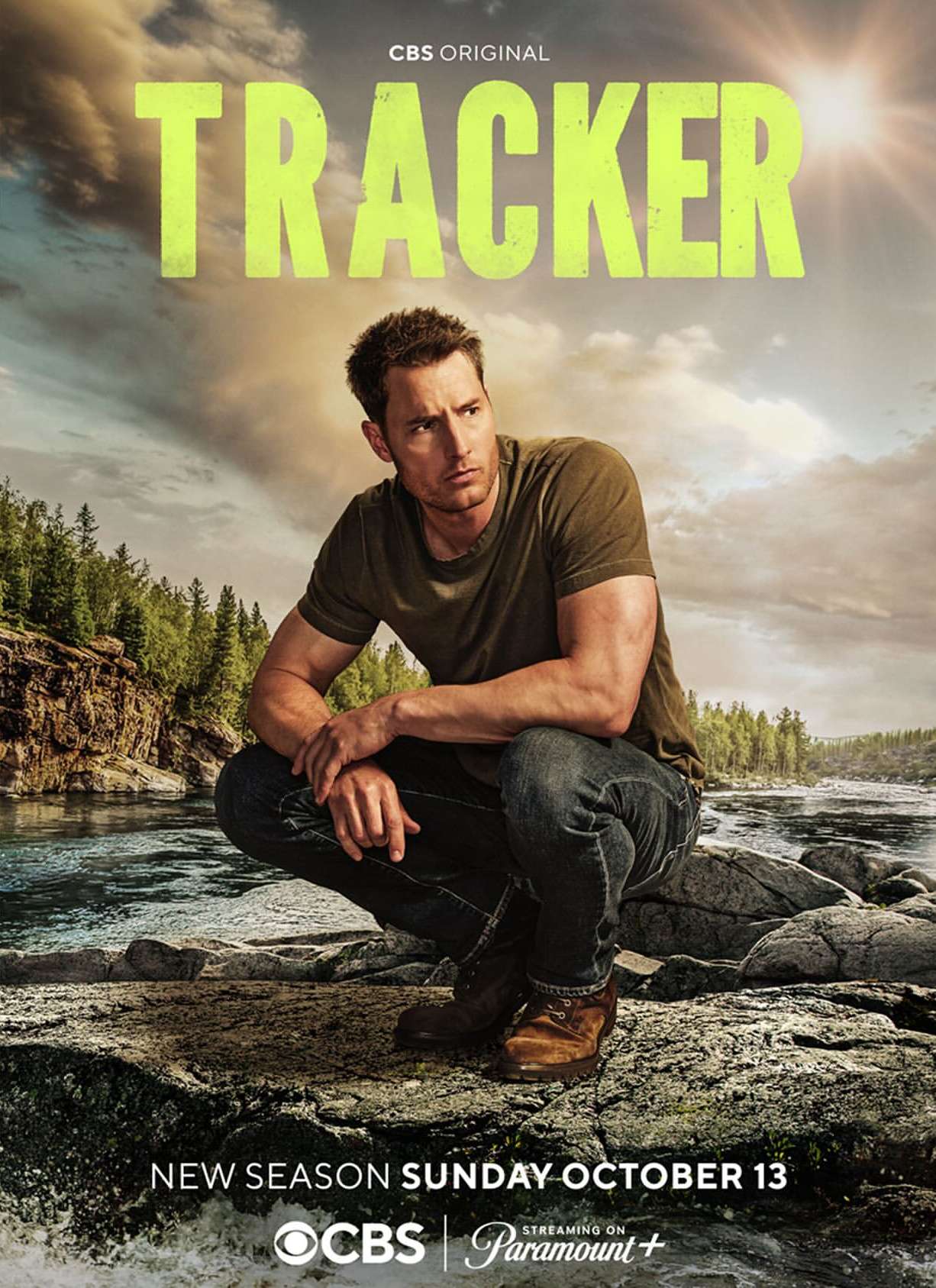 Actor Justin Hartley in a promo image for the new CBS series Tracker, crouching outdoors on a rock with a river backdrop.