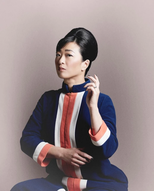 Adult woman with dark hair in retro style posing indoors wearing a blue and orange dress in a portrait-style photograph.