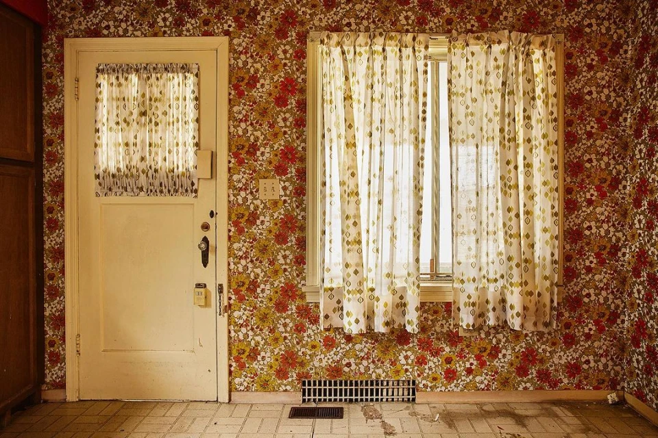 Interior shot of a room featuring floral wallpaper, a white door with a sheer curtained window, and a window with similar curtains.