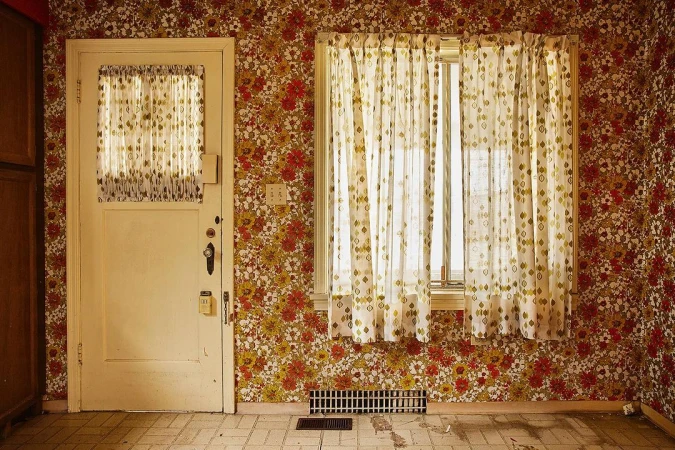 Interior shot of a room featuring floral wallpaper, a white door with a sheer curtained window, and a window with similar curtains.
