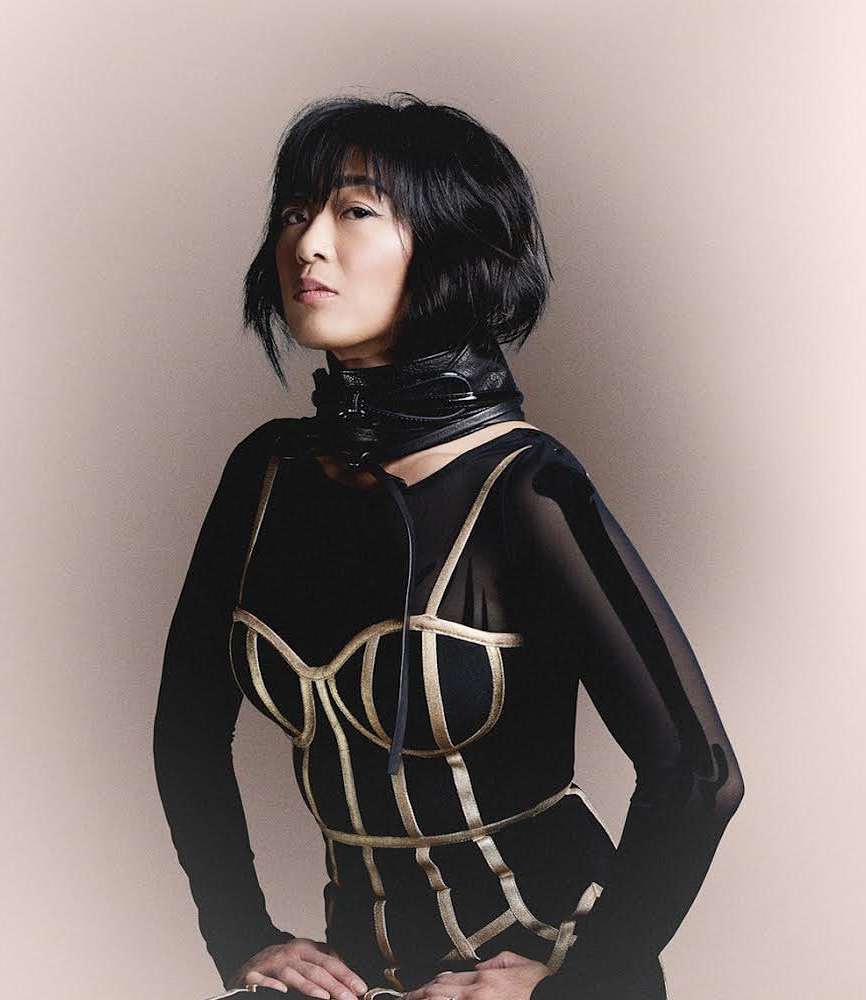 Portrait of an Asian woman in a fashion-forward black and gold bustier with a large black collar, posing in front of a neutral wall.