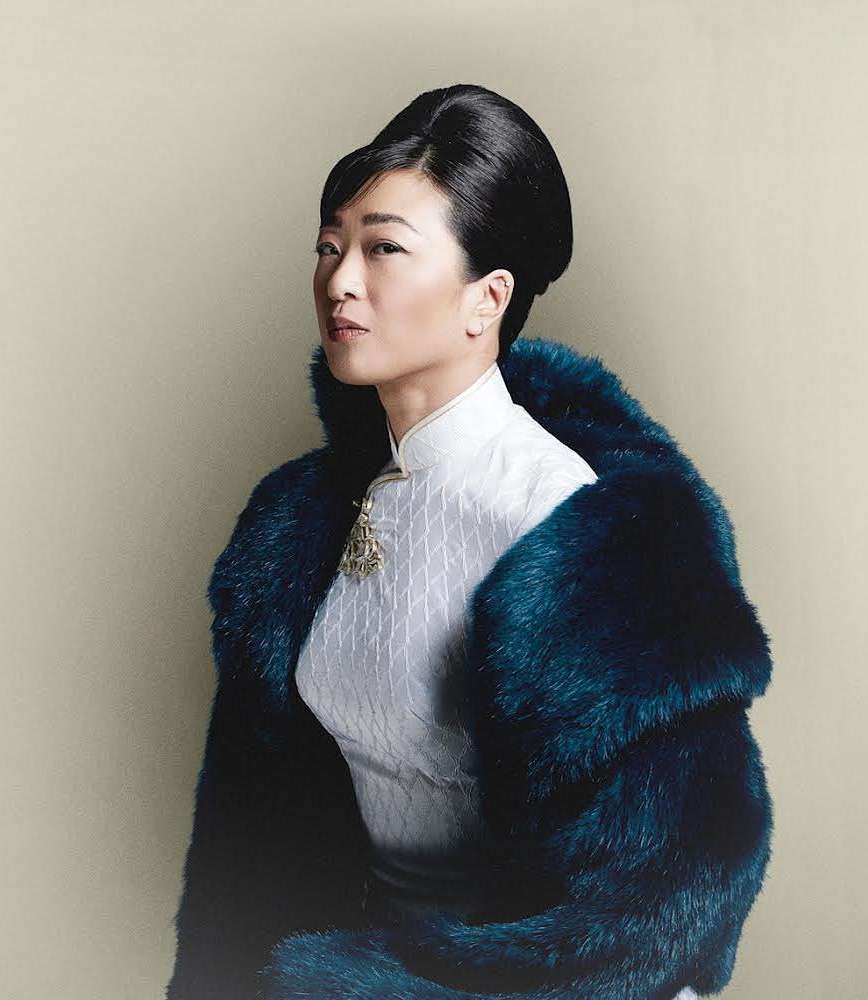 Elegant Asian woman posing indoors wearing traditional white patterned dress and oversized teal faux fur coat with decorative brooch.