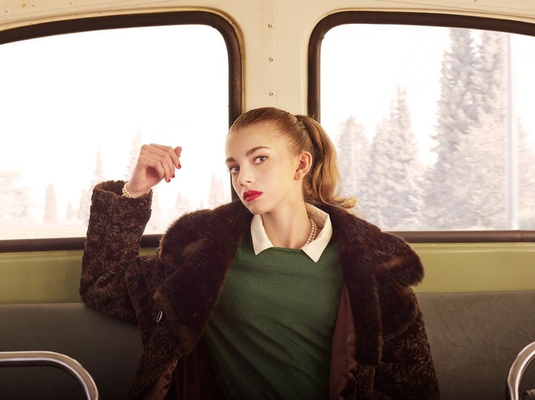 Portrait of a young woman in a vintage style, wearing a dark fur coat and green dress while sitting on a vehicle with large windows.