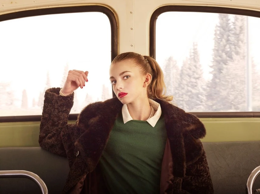 Portrait of a young woman in a vintage style, wearing a dark fur coat and green dress while sitting on a vehicle with large windows.