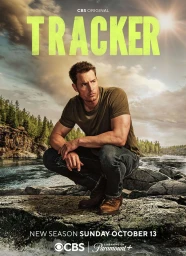 Justin Hartley crouching on rocky riverbank wearing dark t-shirt and jeans with river and forested hills behind