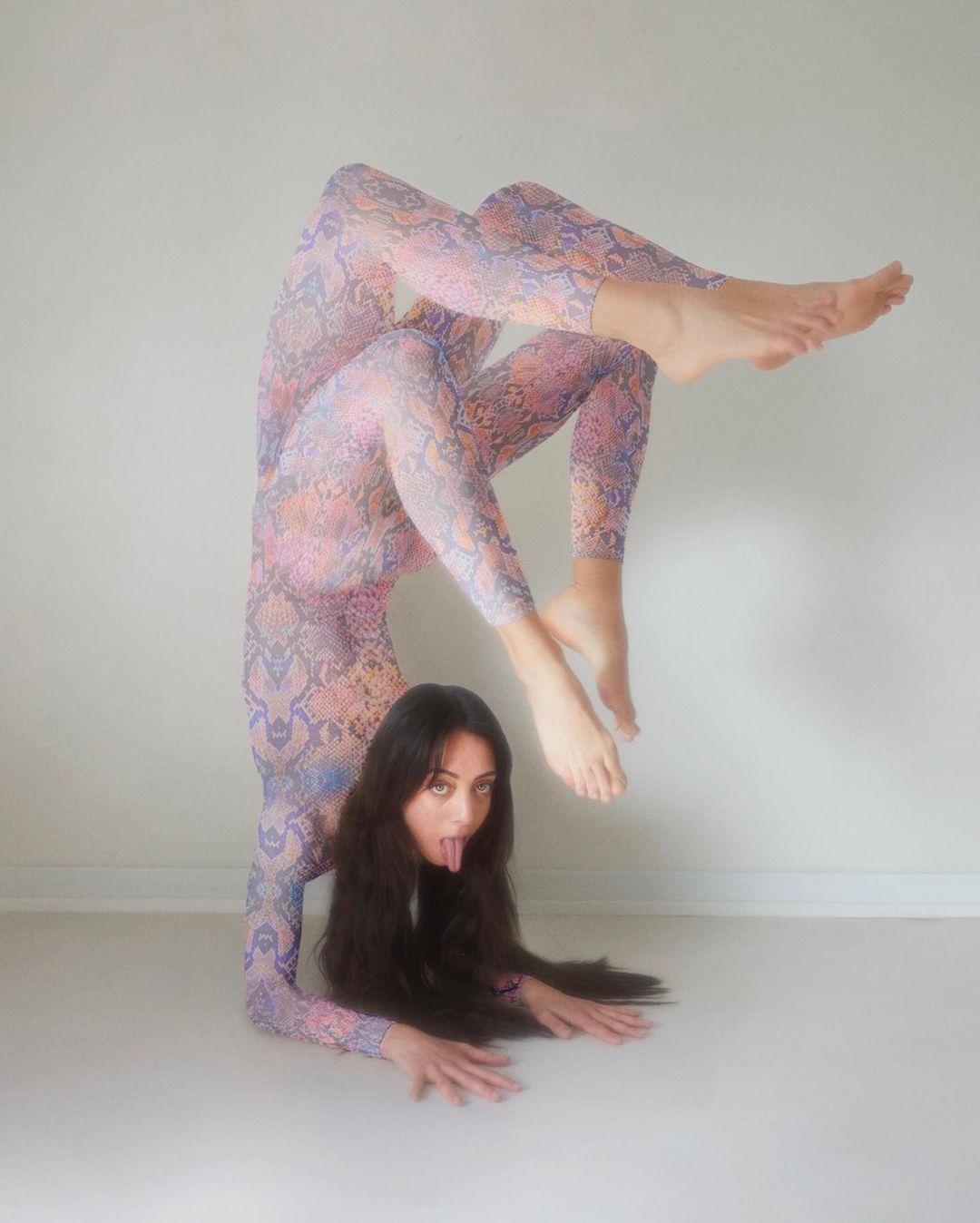 A woman with long dark hair wearing a patterned bodysuit performs a handstand while sticking out her tongue against a white backdrop.