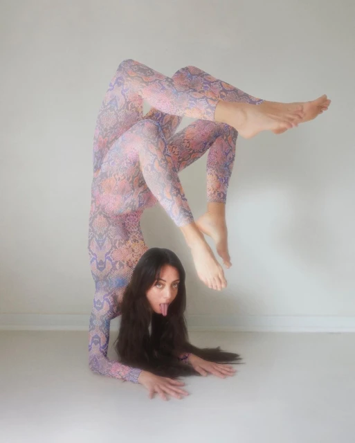 A woman with long dark hair wearing a patterned bodysuit performs a handstand while sticking out her tongue against a white backdrop.