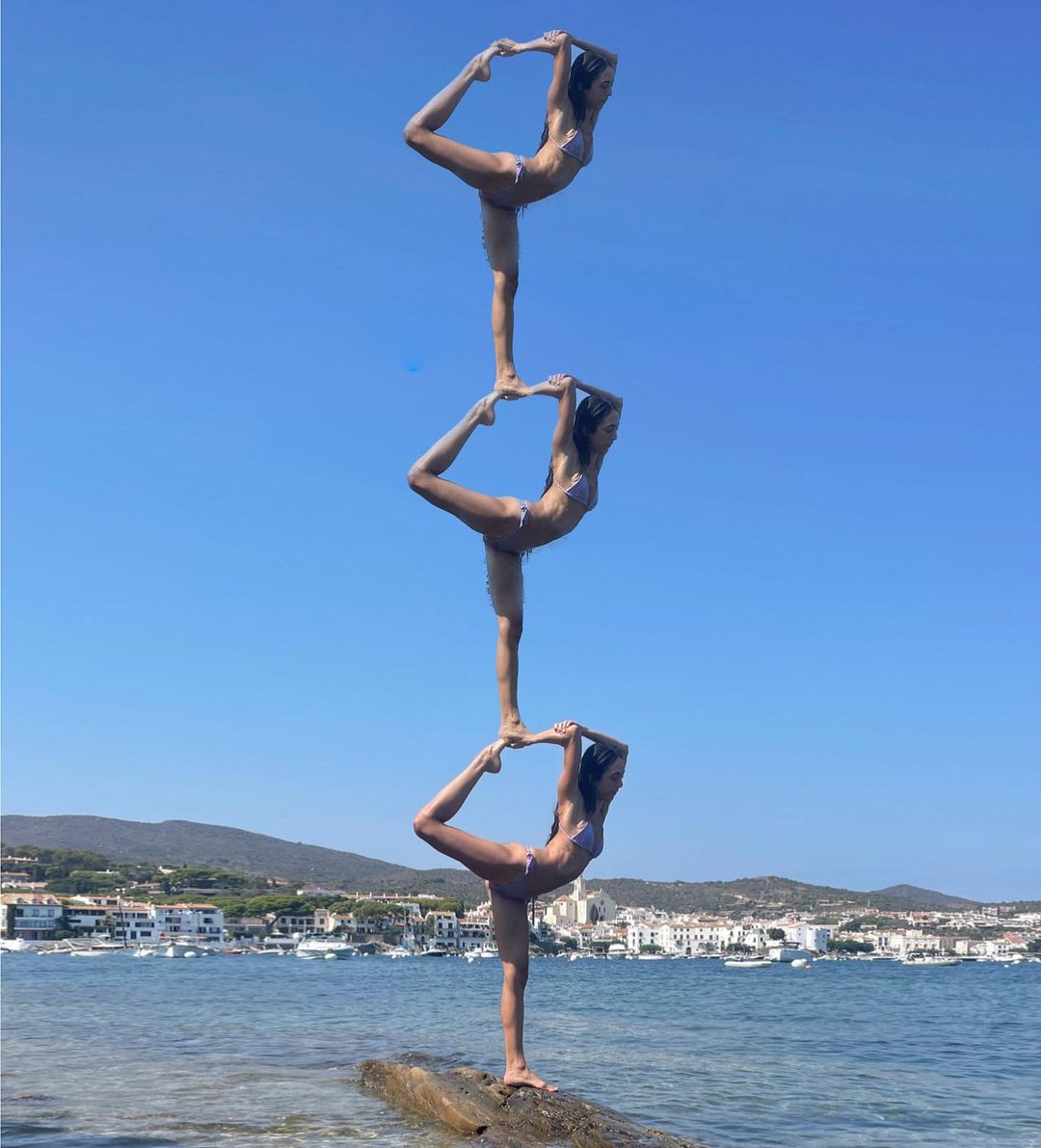 Three adult females stacked on each other in yoga poses in bikinis on a rock near the ocean with a clear blue sky, performing a balance act.