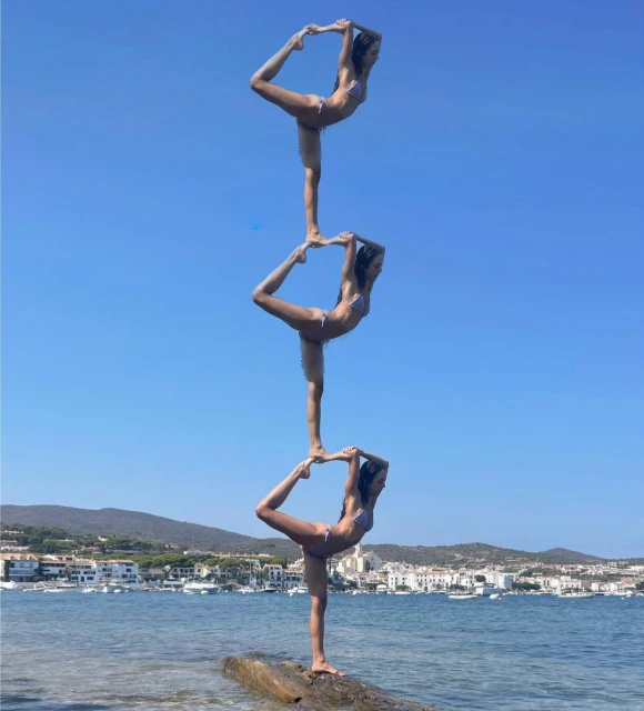 Three adult females stacked on each other in yoga poses in bikinis on a rock near the ocean with a clear blue sky, performing a balance act.