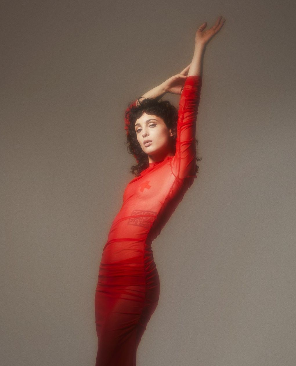 Portrait of a woman in a red, sheer dress with her arms raised, posing against a neutral background, fashion-forward and elegant.