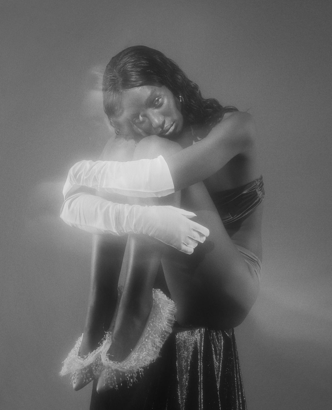 Monochrome portrait of a Black woman in a strapless top, seated on a draped platform, wearing tulle gloves and shoes, embracing her legs.