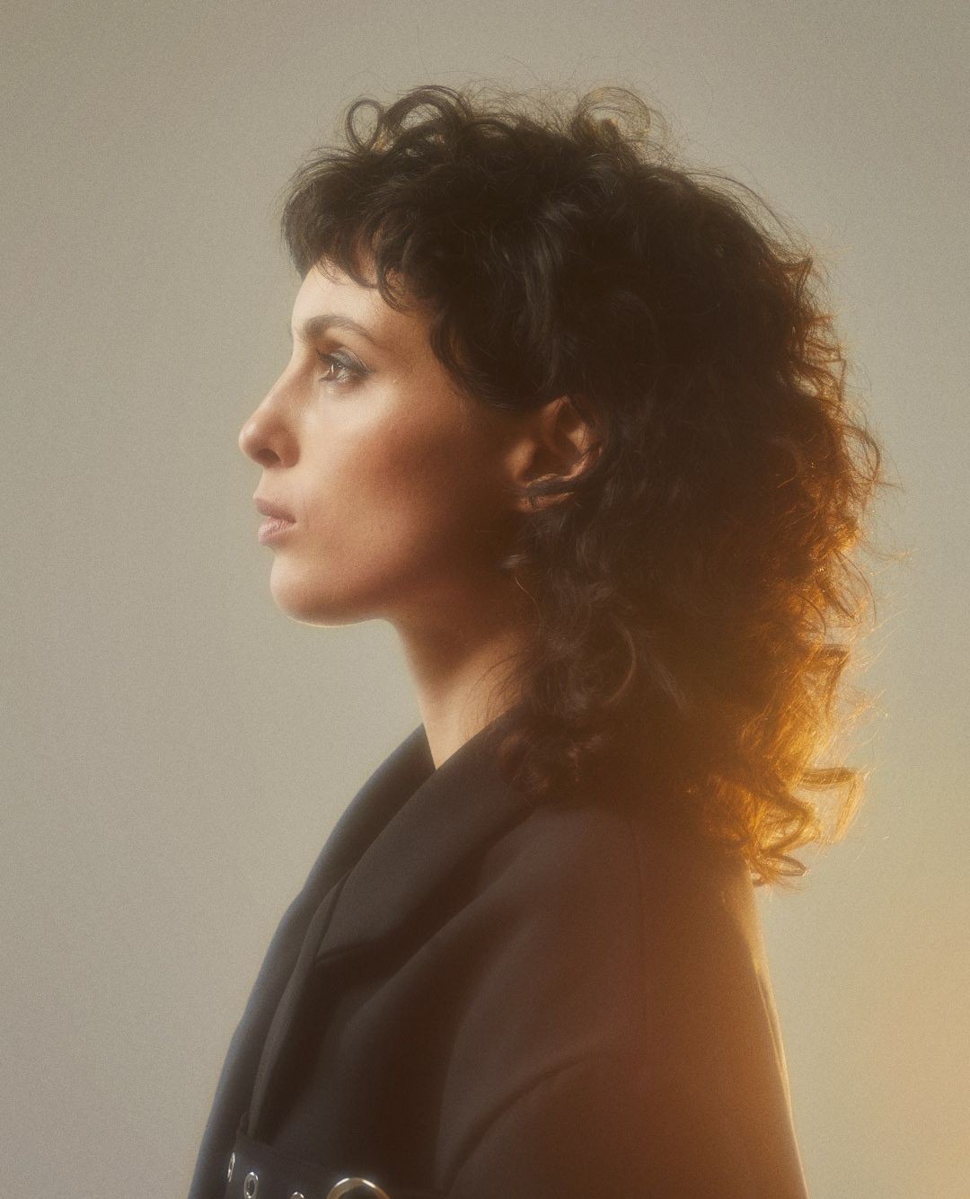 Portrait of an adult female with curly dark hair, wearing a black top with silver grommets, in a profile shot against a neutral backdrop.