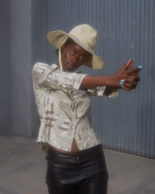 Stylish woman in hat and patterned shirt strikes a pose outdoors, aiming fingers in a gun-like gesture against a gray corrugated background.