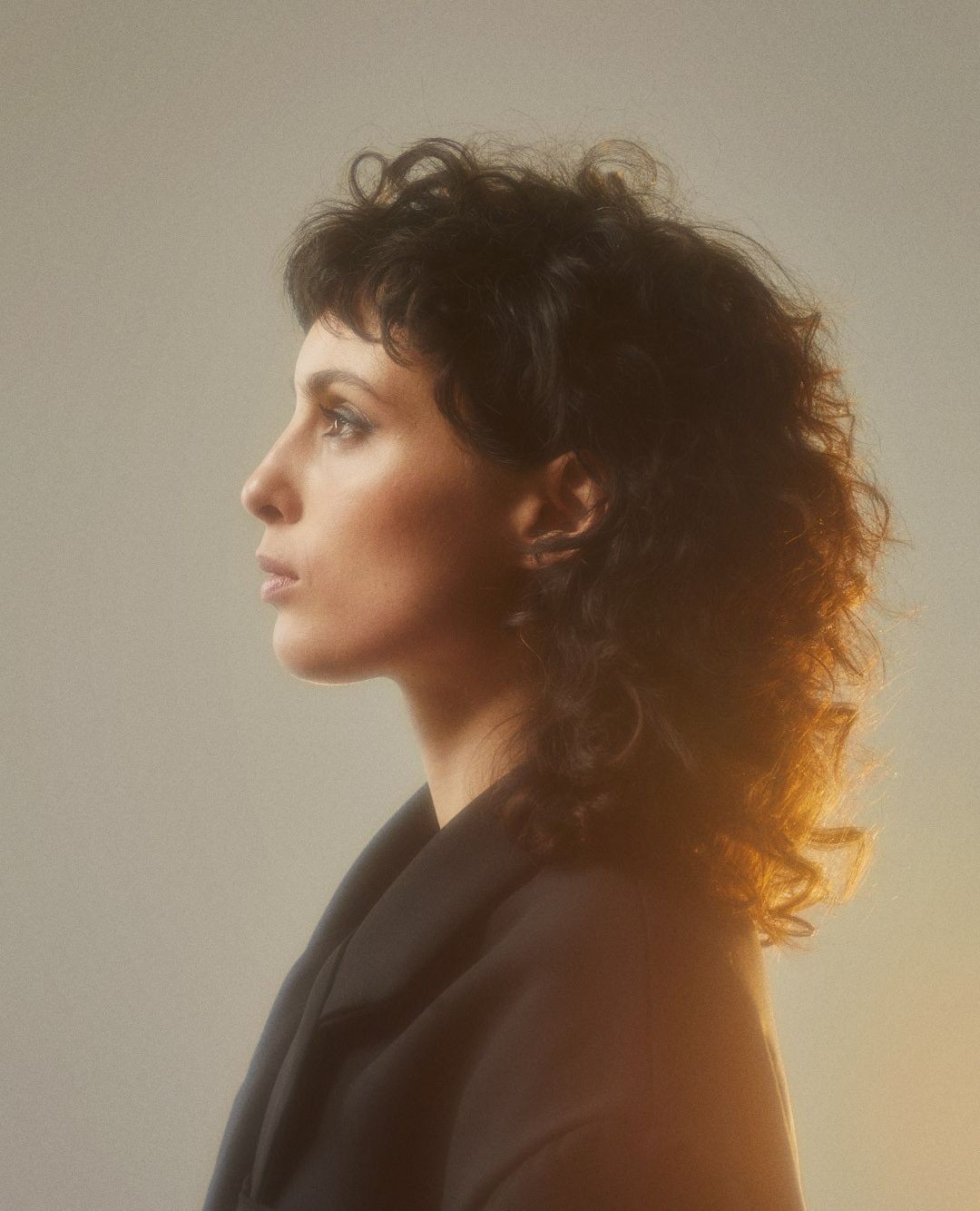 Portrait of a brunette woman with curly hair in profile against a soft gray background, wearing a dark blazer in a photo studio.