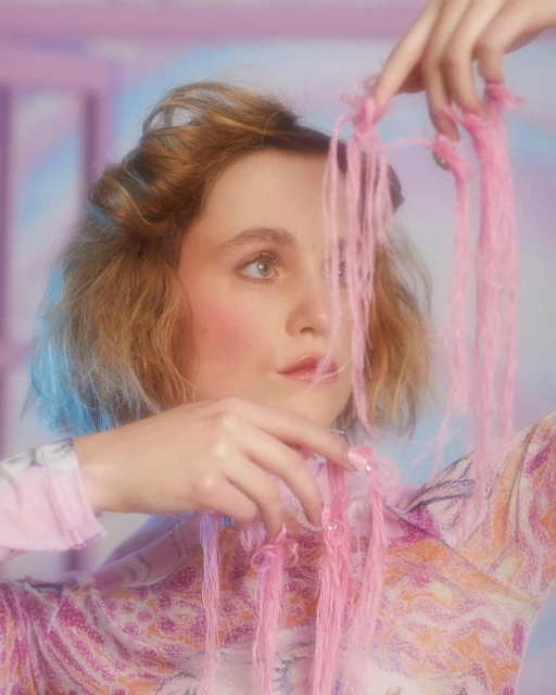 A young adult female with blonde hair and pink blush holding pink threads indoors with a soft blue and pink background.