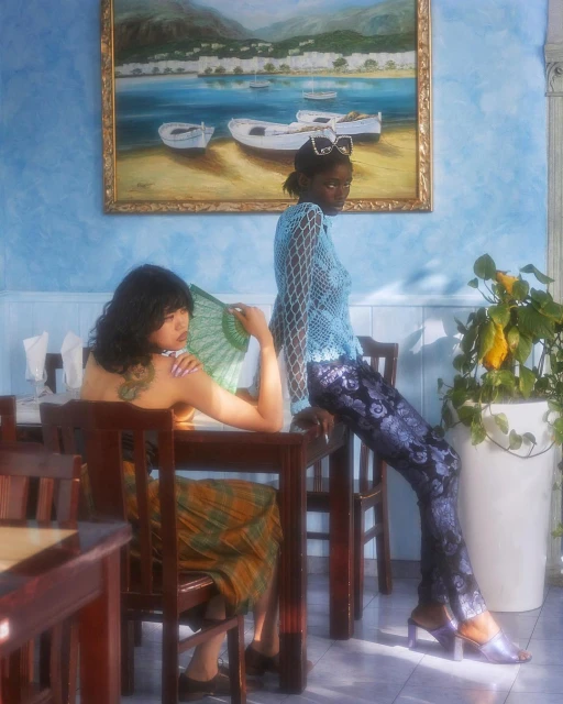Two women in a restaurant setting, one sitting at a table with a fan, the other standing by the table in an airy blue top.