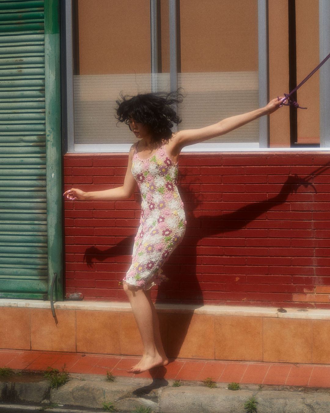 Barefoot adult female in floral dress leaps in an urban setting with a green door and brick wall in the background on a sunny day.