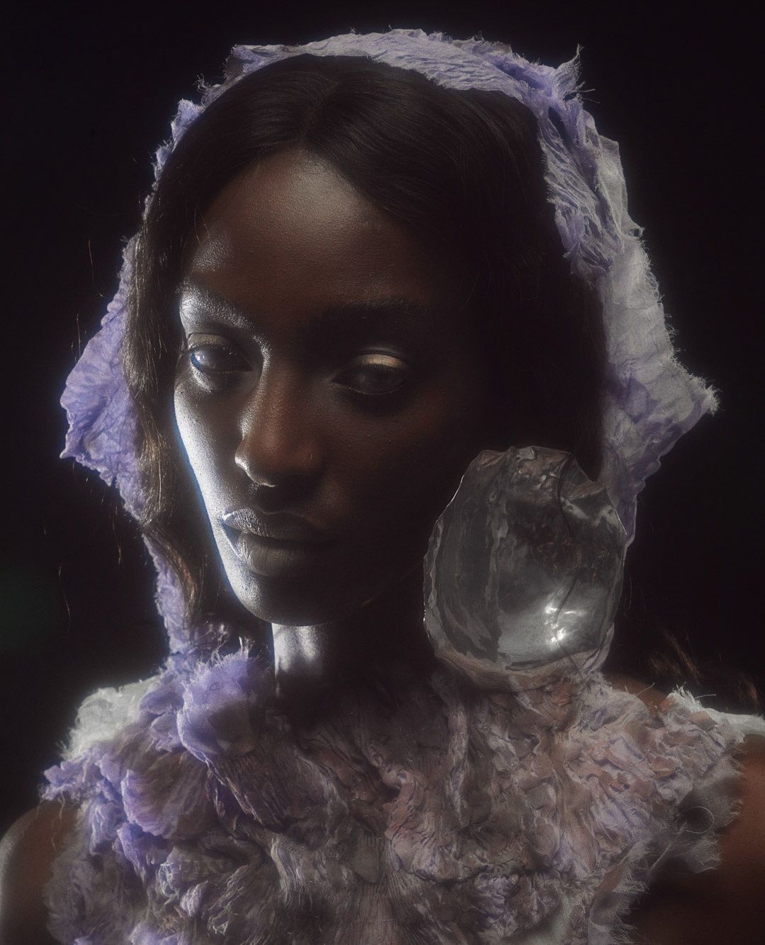 Portrait of an adult female model with dark skin wearing a ruffled collar and hood, both in light purple, posing against a dark backdrop.