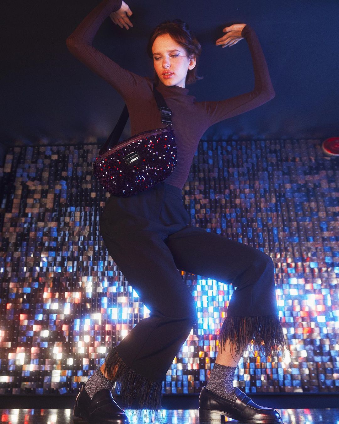 Stylish adult woman poses dynamically in flared black fringe trousers, shiny socks and loafers, wearing a sparkly crossbody bag against a sparkly wall.