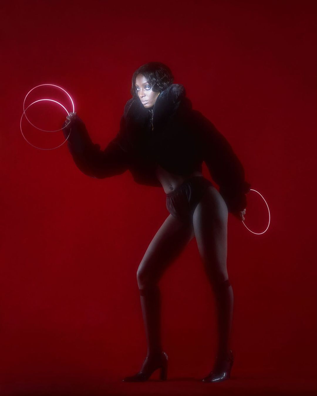 A woman poses in a cropped black jacket and heels against a red backdrop, holding two glowing ring-shaped lights for a fashion or editorial shoot.