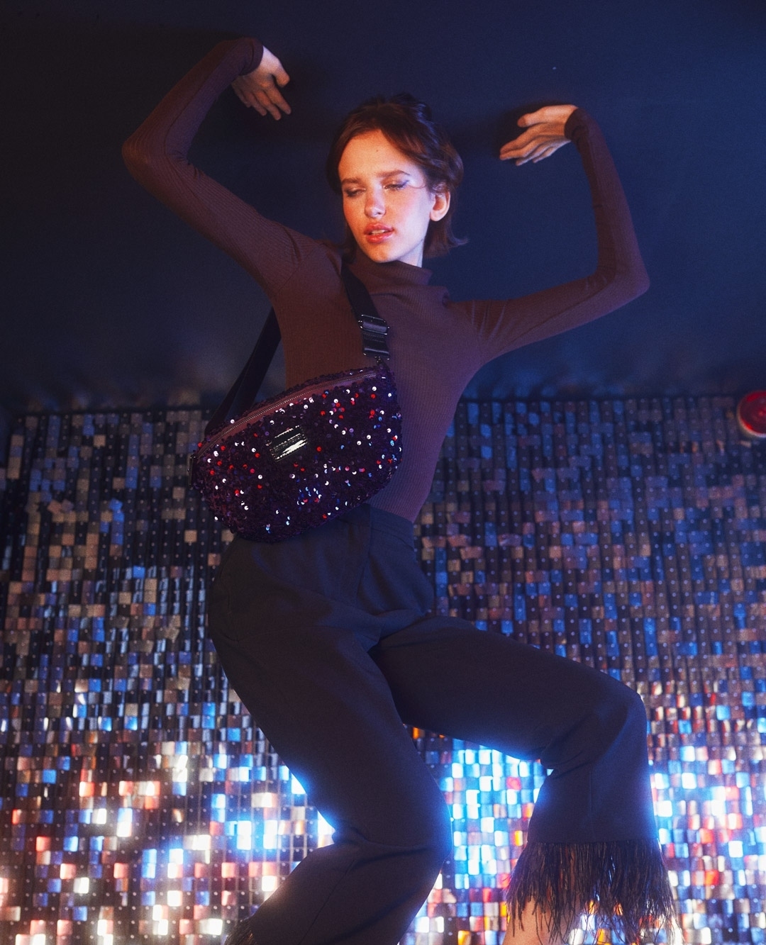 Woman in a long-sleeve top and fringe pants poses with a burgundy, sequined crossbody bag against a dark, shimmering background.