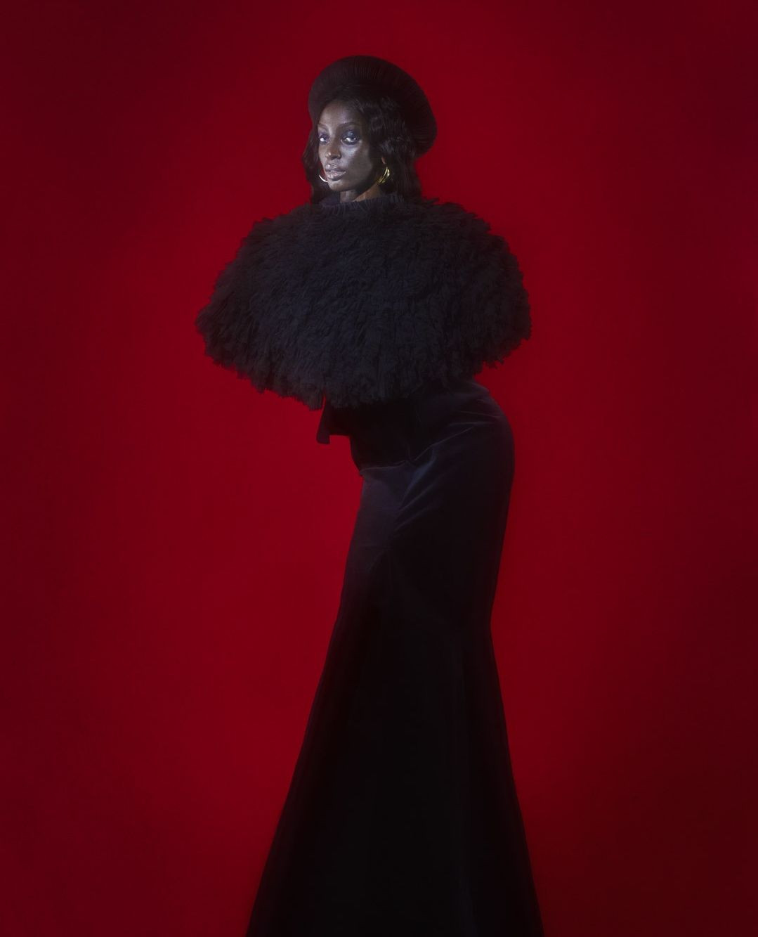 Elegant Black woman in black velvet gown and fur-like bolero poses against a red studio background, showcasing dramatic fashion.