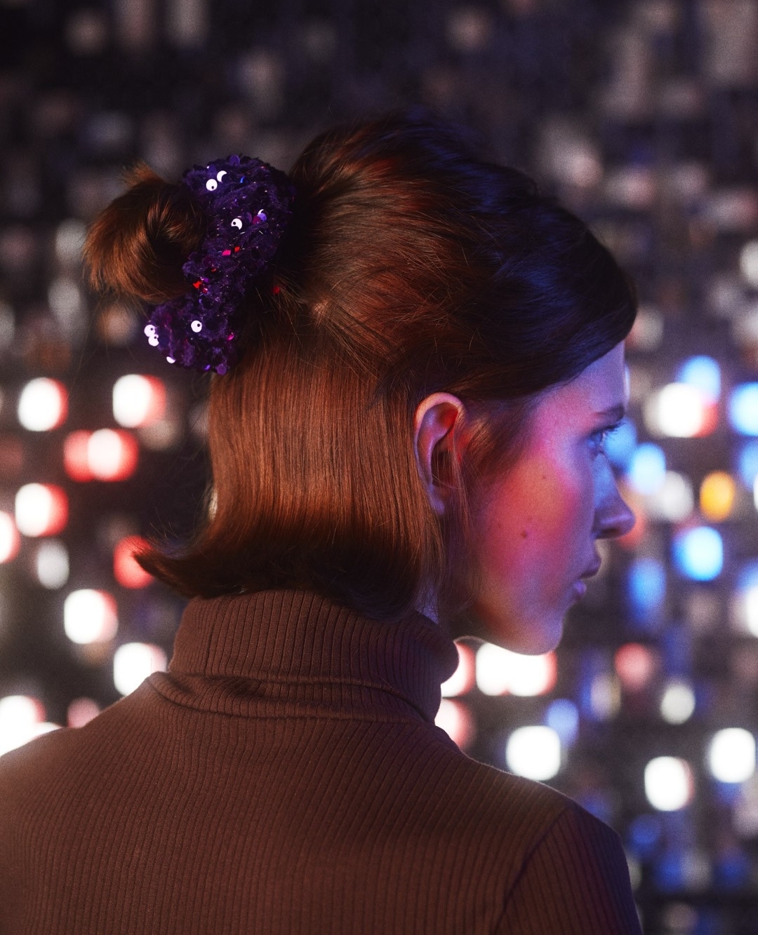 Woman with bob hairstyle wearing black turtleneck and purple sequin hair accessory in front of blurred bokeh lights.