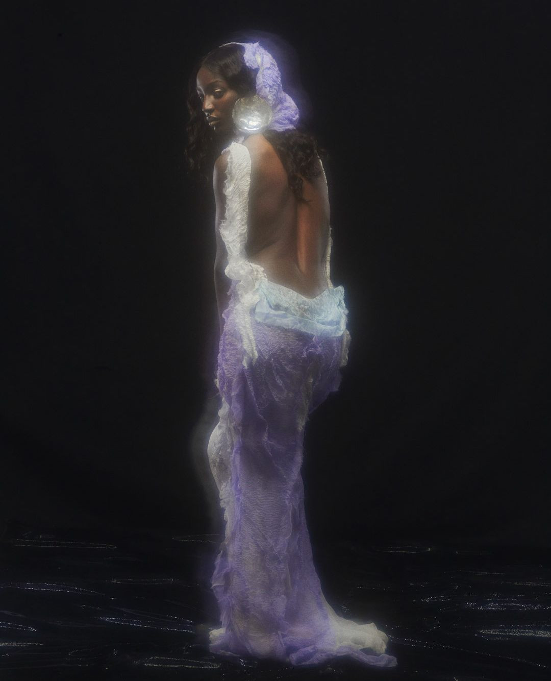A person with dark hair wearing a light purple dress and head covering in a dark environment poses with their back to the camera.