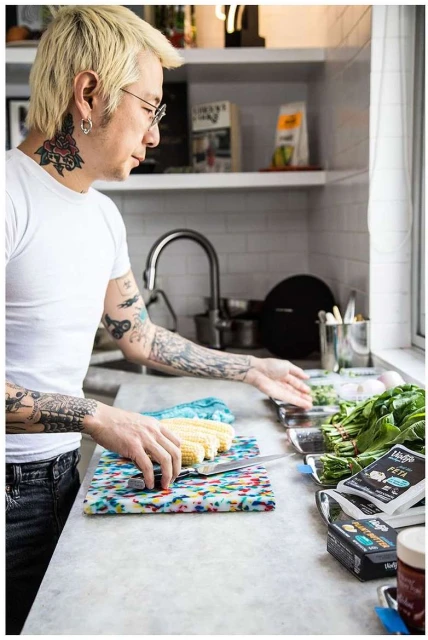Asian man with tattoos prepares vegetables in a kitchen; chopping corn on a colorful cutting board near spinach and plant-based feta.