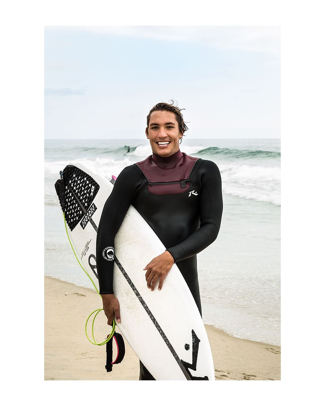 Smiling surfer in wetsuit poses with surfboard on sandy beach with ocean waves. Summer beach scene with clear sky and blue water.