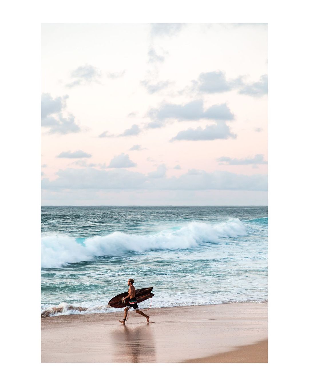 A surfer runs along a sandy beach carrying a surfboard, with ocean waves and a sky filled with pastel clouds in the background.