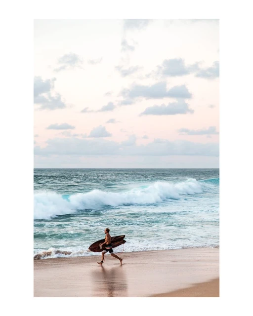 A surfer runs along a sandy beach carrying a surfboard, with ocean waves and a sky filled with pastel clouds in the background.