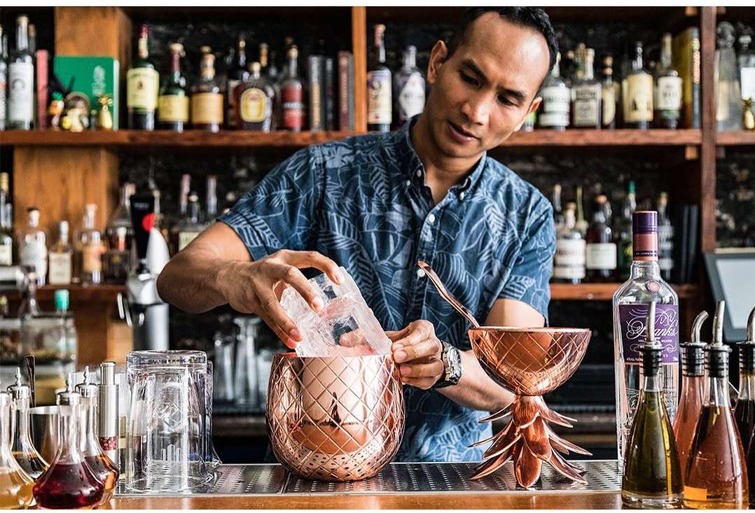Adult male bartender makes cocktails at bar, pouring ice into pineapple-shaped copper mug with bar shelf of alcohol in background.
