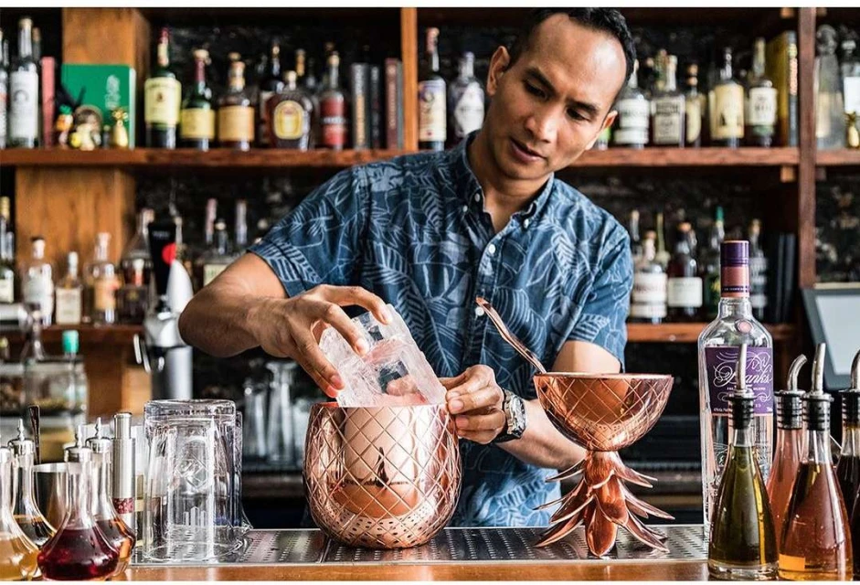 Adult male bartender makes cocktails at bar, pouring ice into pineapple-shaped copper mug with bar shelf of alcohol in background.