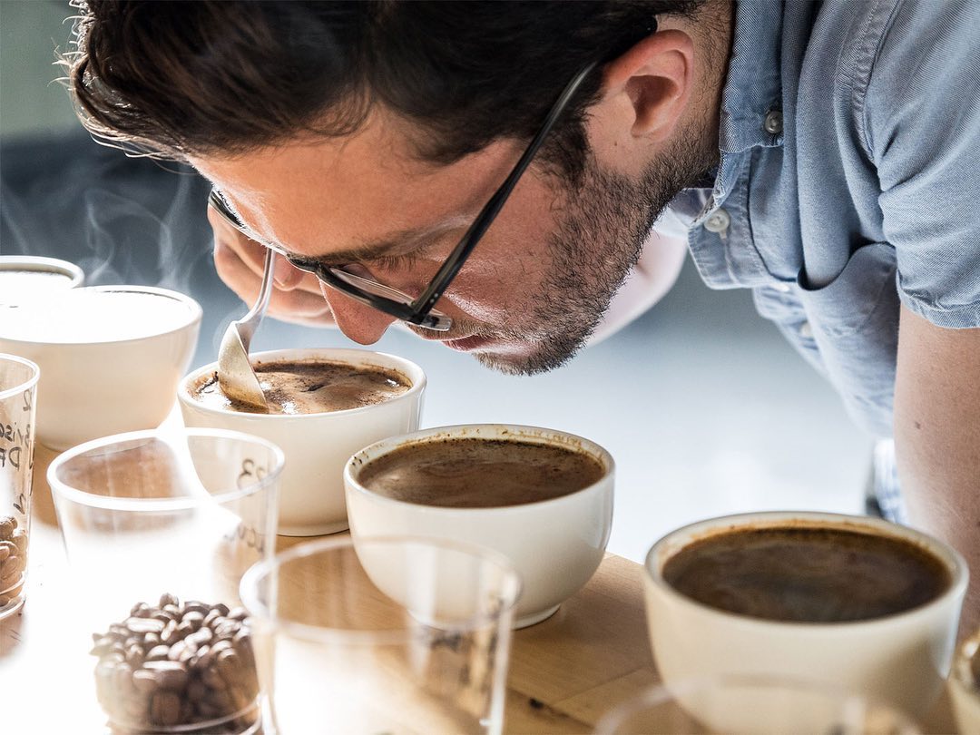 Adult male in eyeglasses leaning over and tasting coffee from a white ceramic bowl, surrounded by coffee beans and drinking glasses.