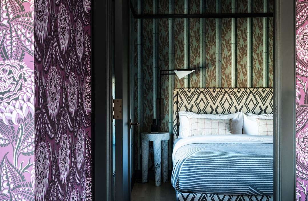 Bedroom interior featuring a bed with striped bedding, patterned headboard, wallpaper, and marble side table viewed through a doorway.