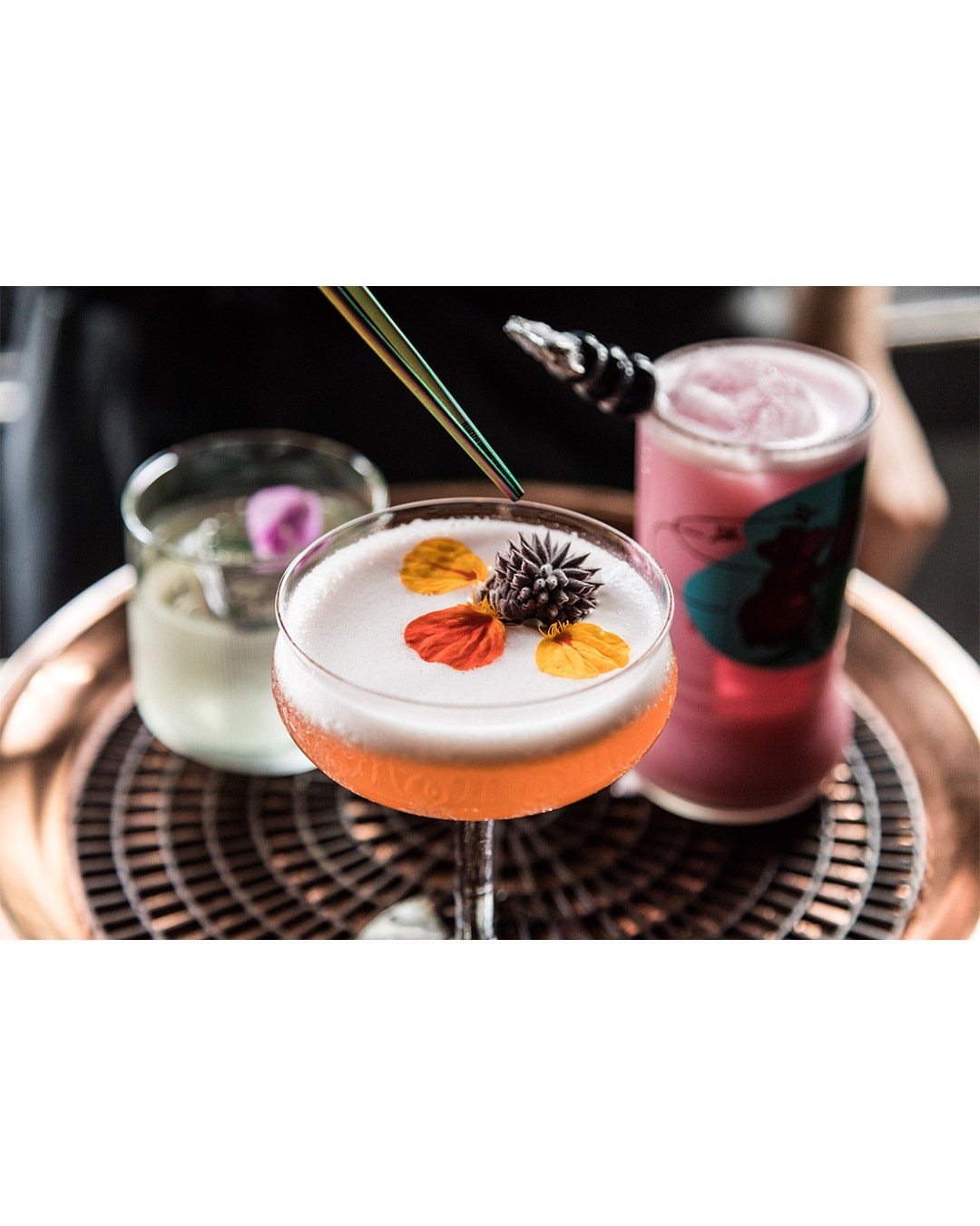 A trio of cocktails featuring an orange cocktail garnished with flower petals, served on a decorative tray with a black background.