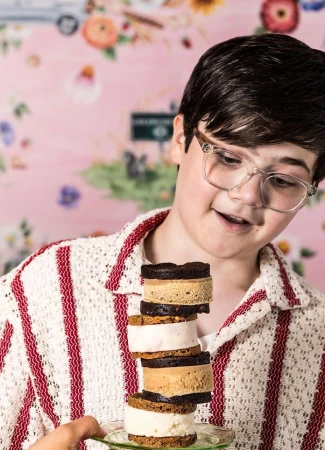 Person holding a tall stack of colorful ice cream sandwiches, with a floral background, wearing clear glasses and a white sweater.