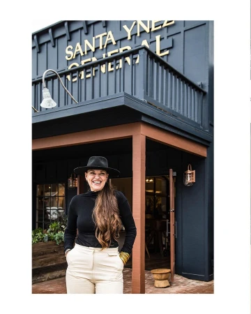 Smiling woman with long brown hair wearing a black hat, black long-sleeve top, and cream trousers standing outside Santa Ynez General store