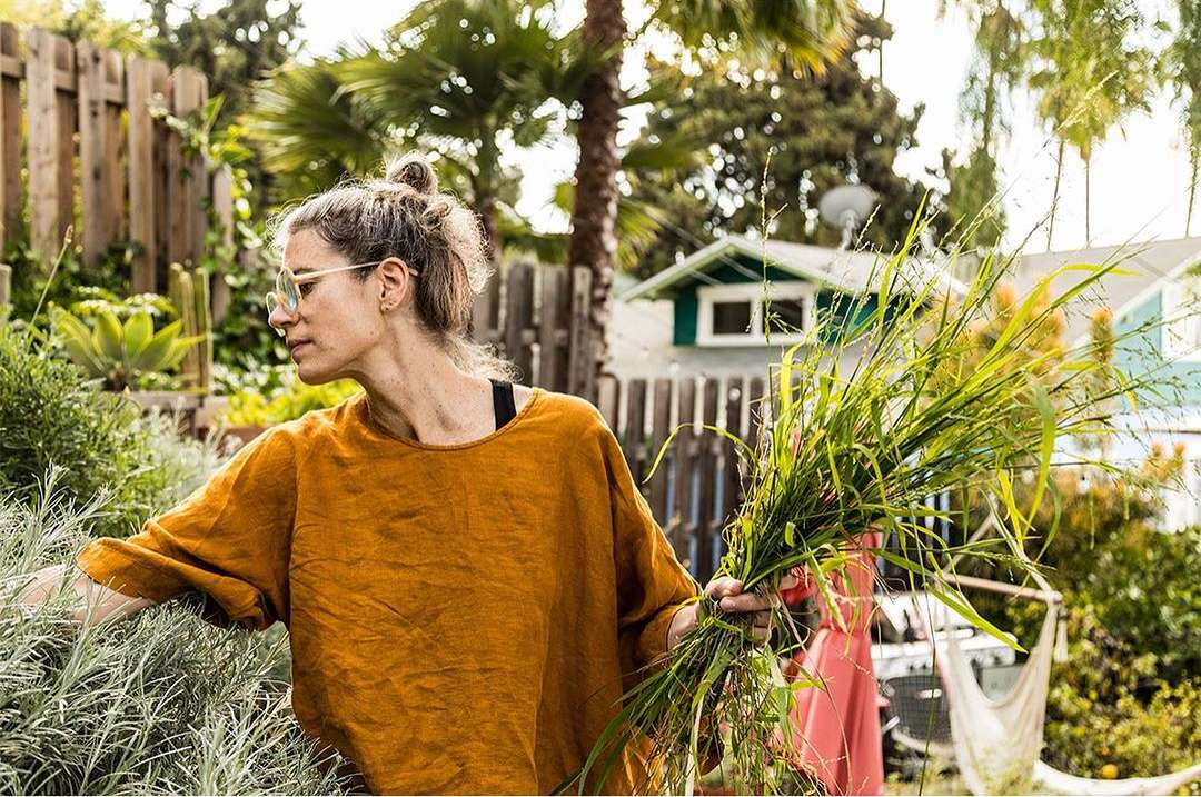 Woman wearing glasses and an orange shirt tends to a garden with herbs and grasses in the backyard of a residential home on a sunny day.