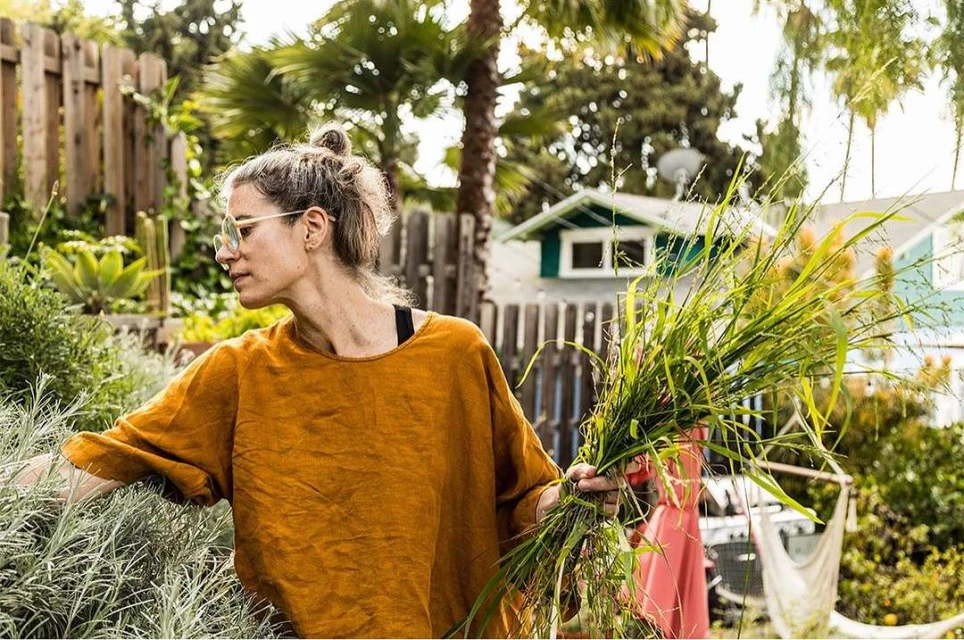 Woman wearing glasses and an orange shirt tends to a garden with herbs and grasses in the backyard of a residential home on a sunny day.